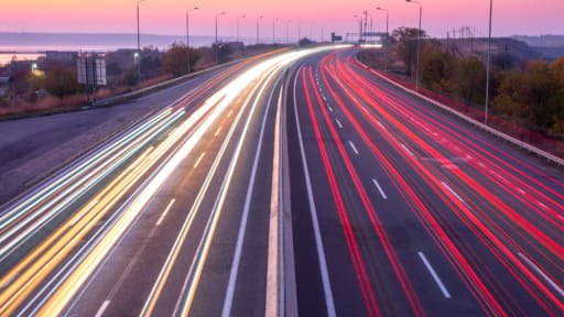Langzeitbelichtung einer Autobahn bei Dämmerung mit leuchtenden Lichtspuren von Fahrzeugen in Weiß und Rot, die eine dynamische Bewegung auf den mehrspurigen Fahrbahnen darstellen.