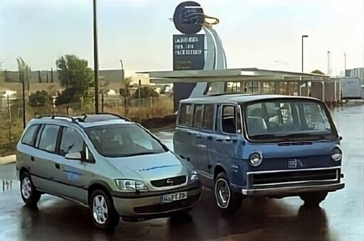 Zwei Fahrzeuge vor einer Wasserstoff-Tankstelle. Links steht ein silberner Opel Zafira, rechts ein blauer GM Electrovan aus dem Jahr 1966. Der Himmel ist bewölkt und die Straße ist nass.