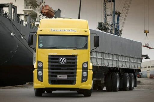 Ein gelber Lastwagen der Marke Volkswagen Constellation steht vor einem Frachtschiff im Hafen. Der LKW hat eine große, silberne Kühlergrill und eine geschlossene Plane auf seinem Anhänger. Im Hintergrund sind Hafenkräne und weitere Frachtschiffe zu sehen.