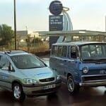 Zwei Fahrzeuge vor einer Wasserstoff-Tankstelle. Links steht ein silberner Opel Zafira, rechts ein blauer GM Electrovan aus dem Jahr 1966. Der Himmel ist bewölkt und die Straße ist nass.